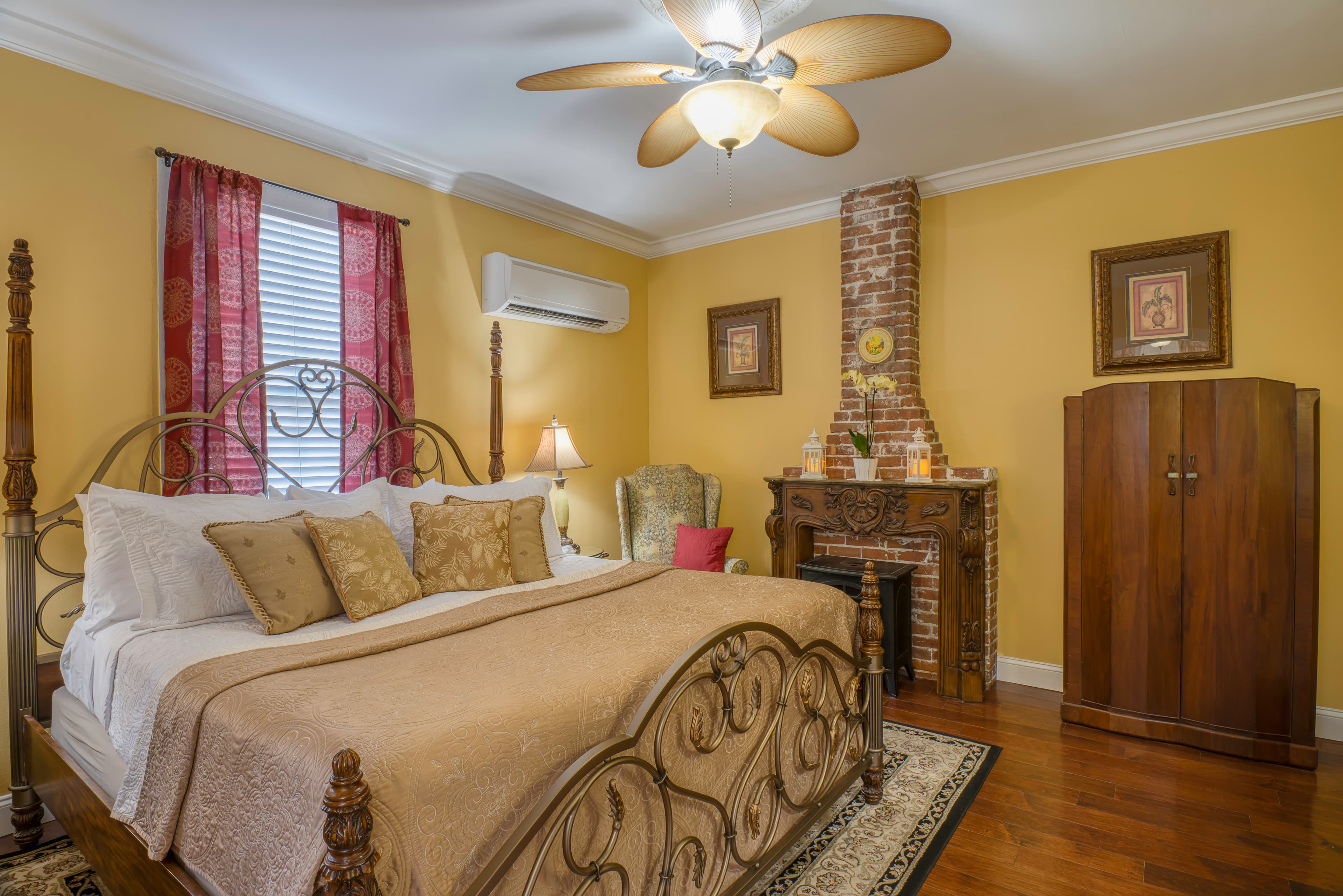 Bedroom in warm yellow and red tones with king brass bed, hardwood floors, electric fireplace, and ceiling fan