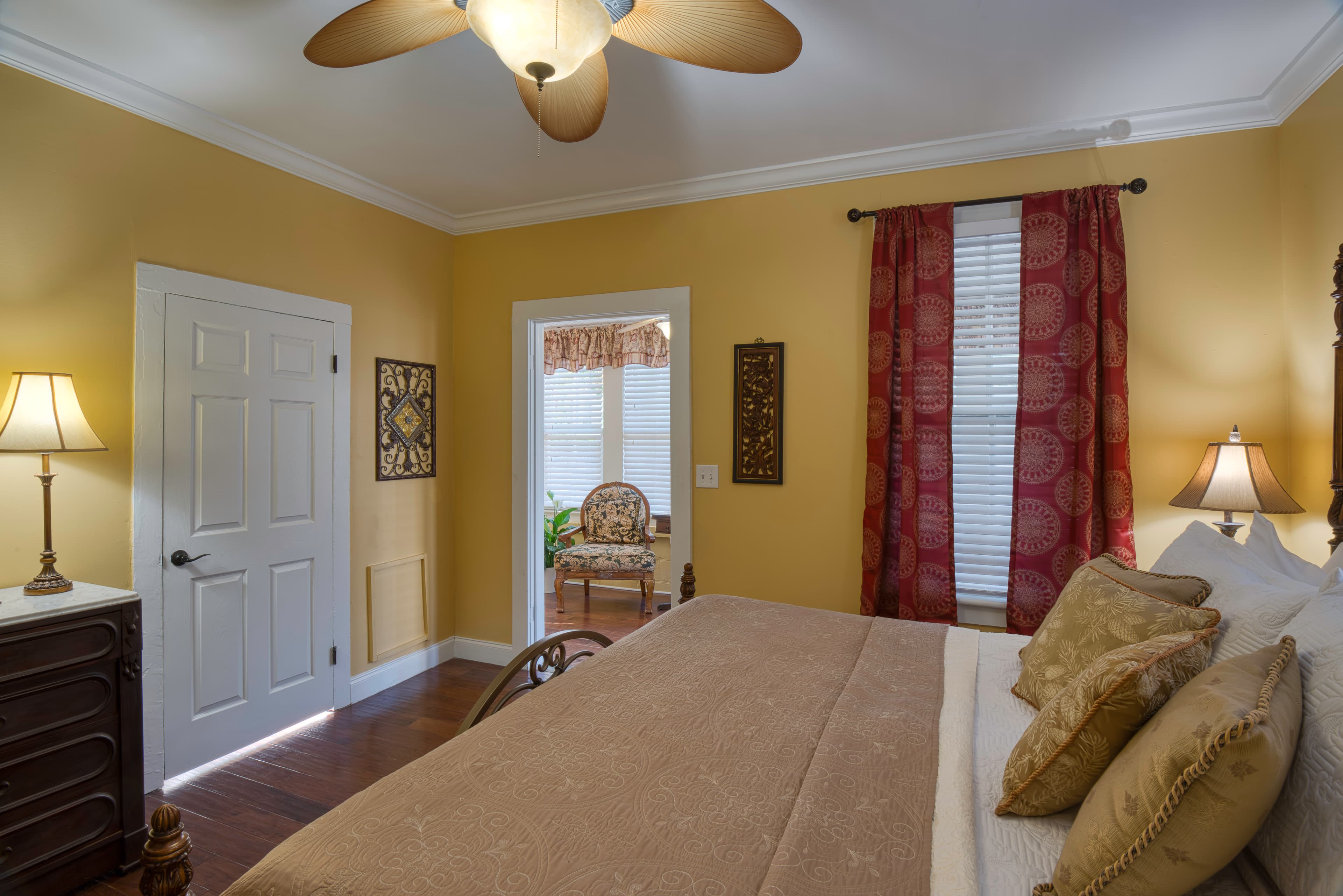 Bedroom with king brass bed, hardwood floors, windows with rich red curtains, doorway to enclosed porch