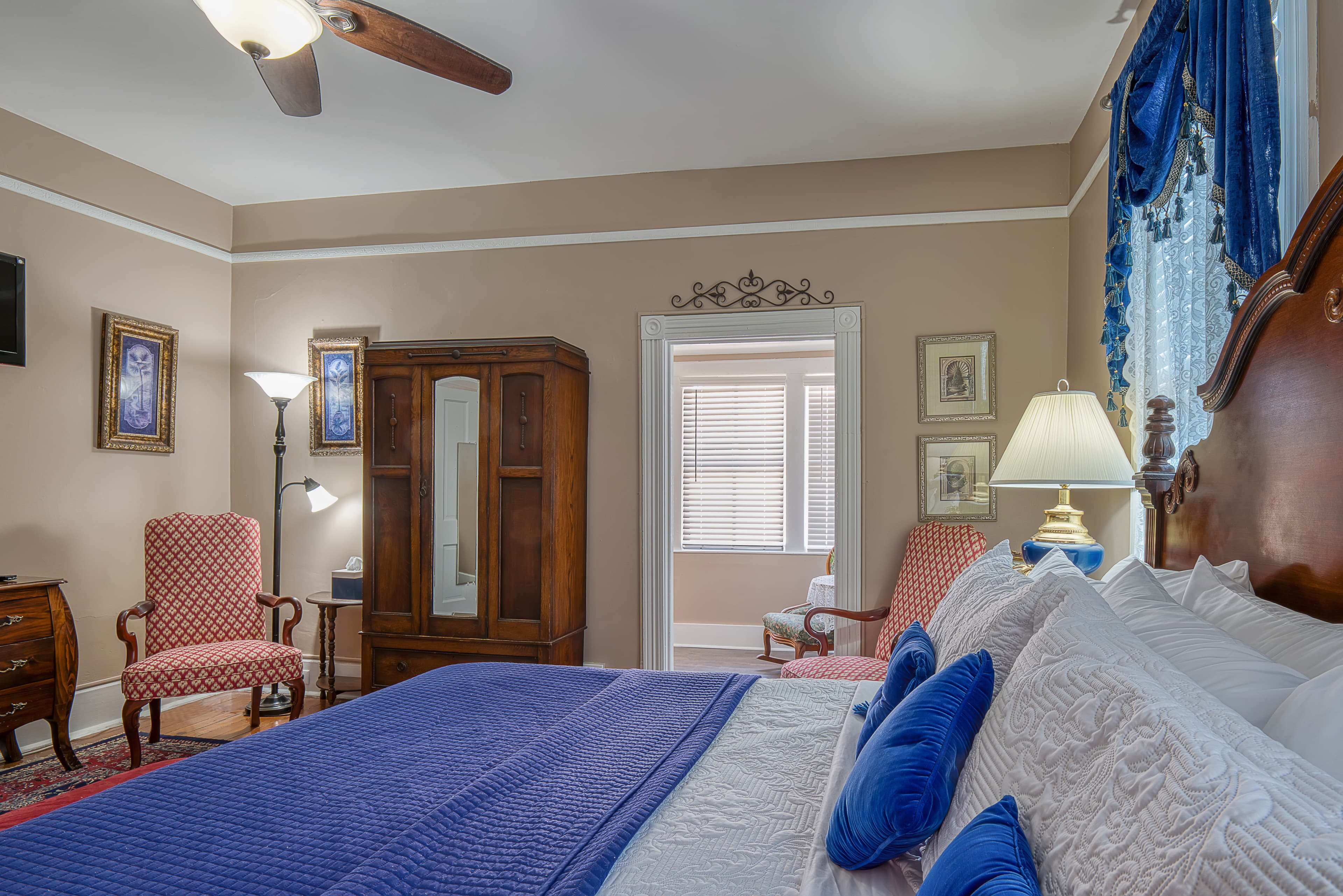 View of bedroom from side of bed in foreground, salmon pink armchairs flanking wooden armoire and doorway leading to enclosed porch