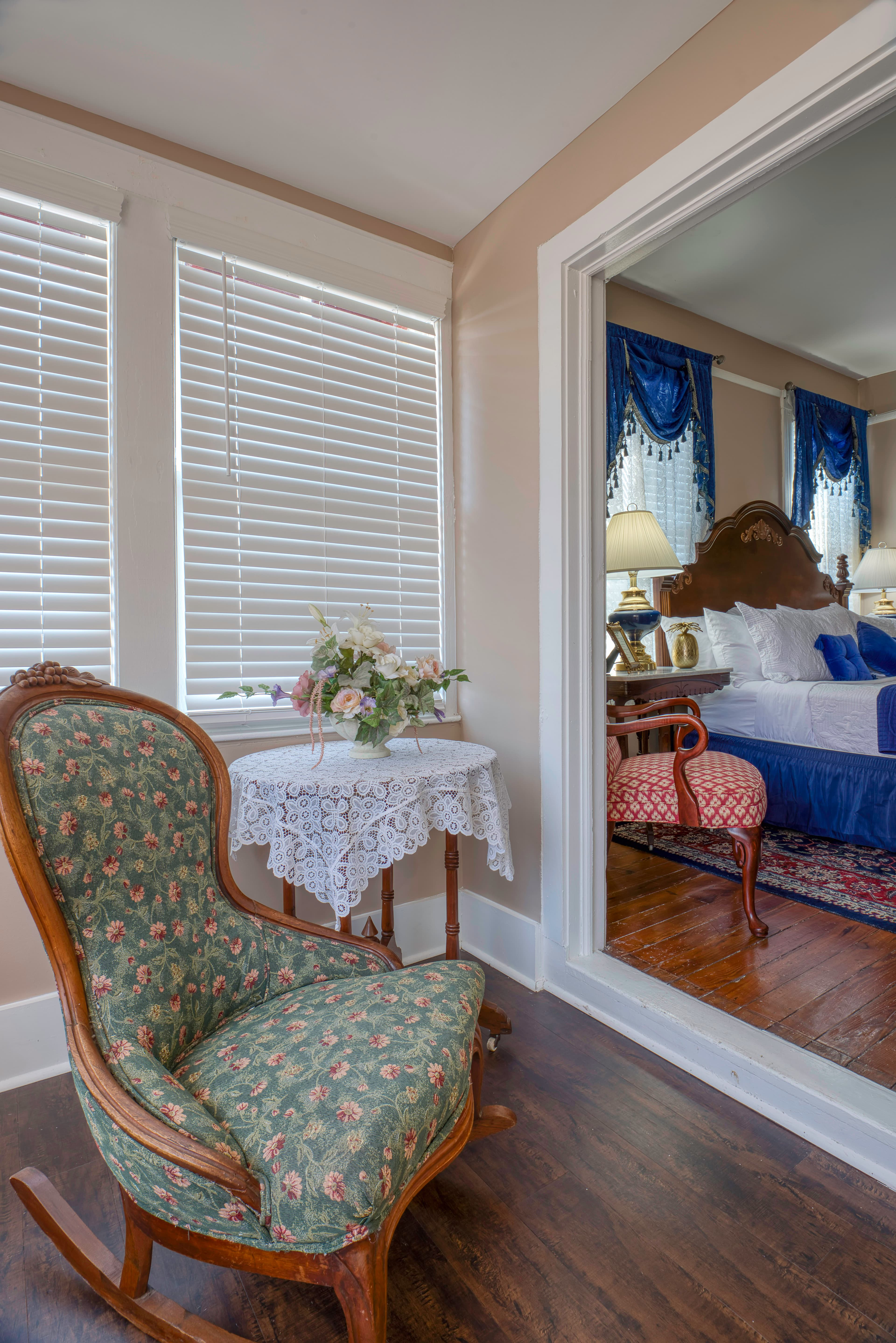 Corner of sitting area on enclosed porch, wooden upholstered rocking chair with round occasional table topped with lace tablecloth and vase of flowers, doorway leading to bedroom