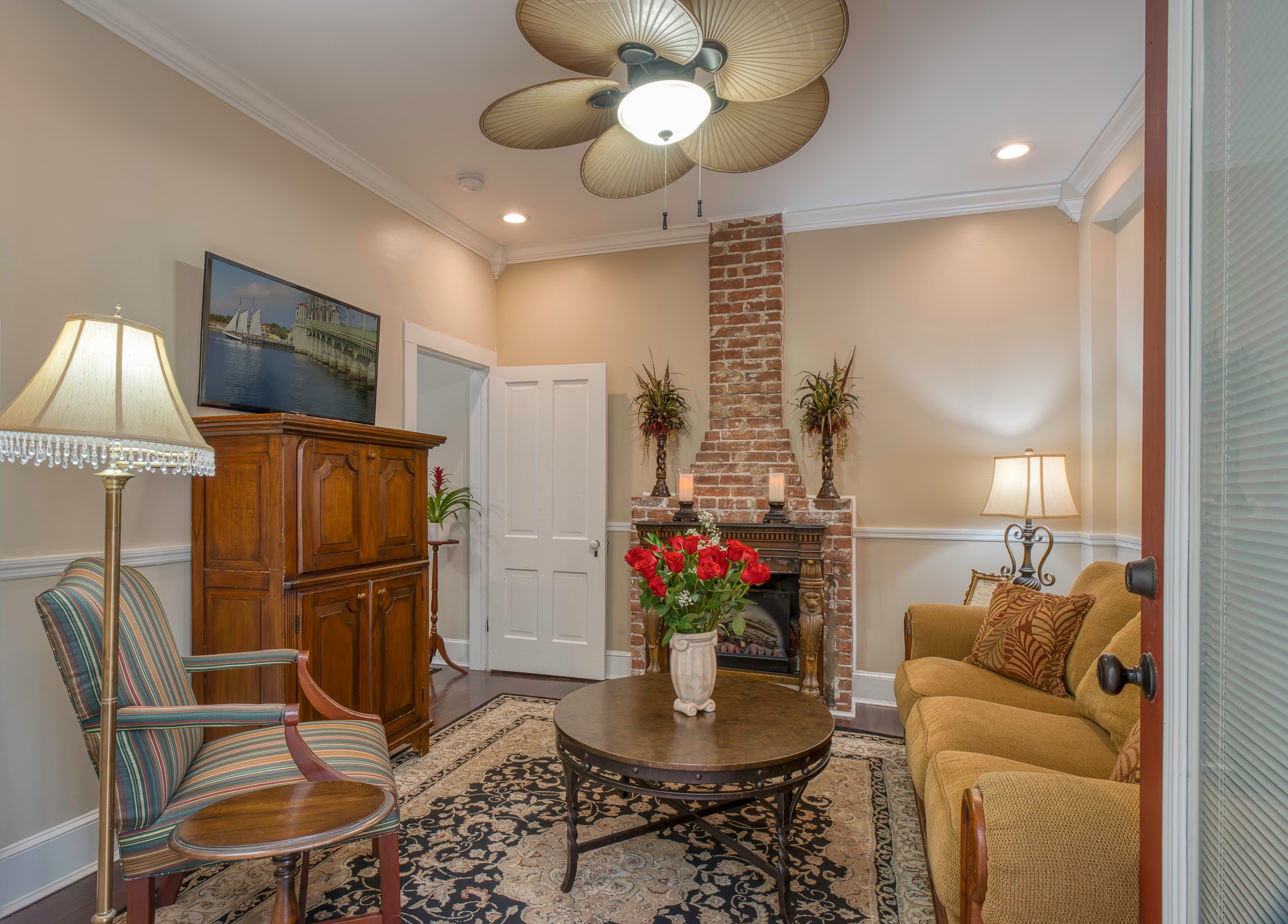 Living room with deep gold sofa bed, round coffee table, comfortable armchair, wooden armoire, wall-mounted flat-screen TV, and fireplace