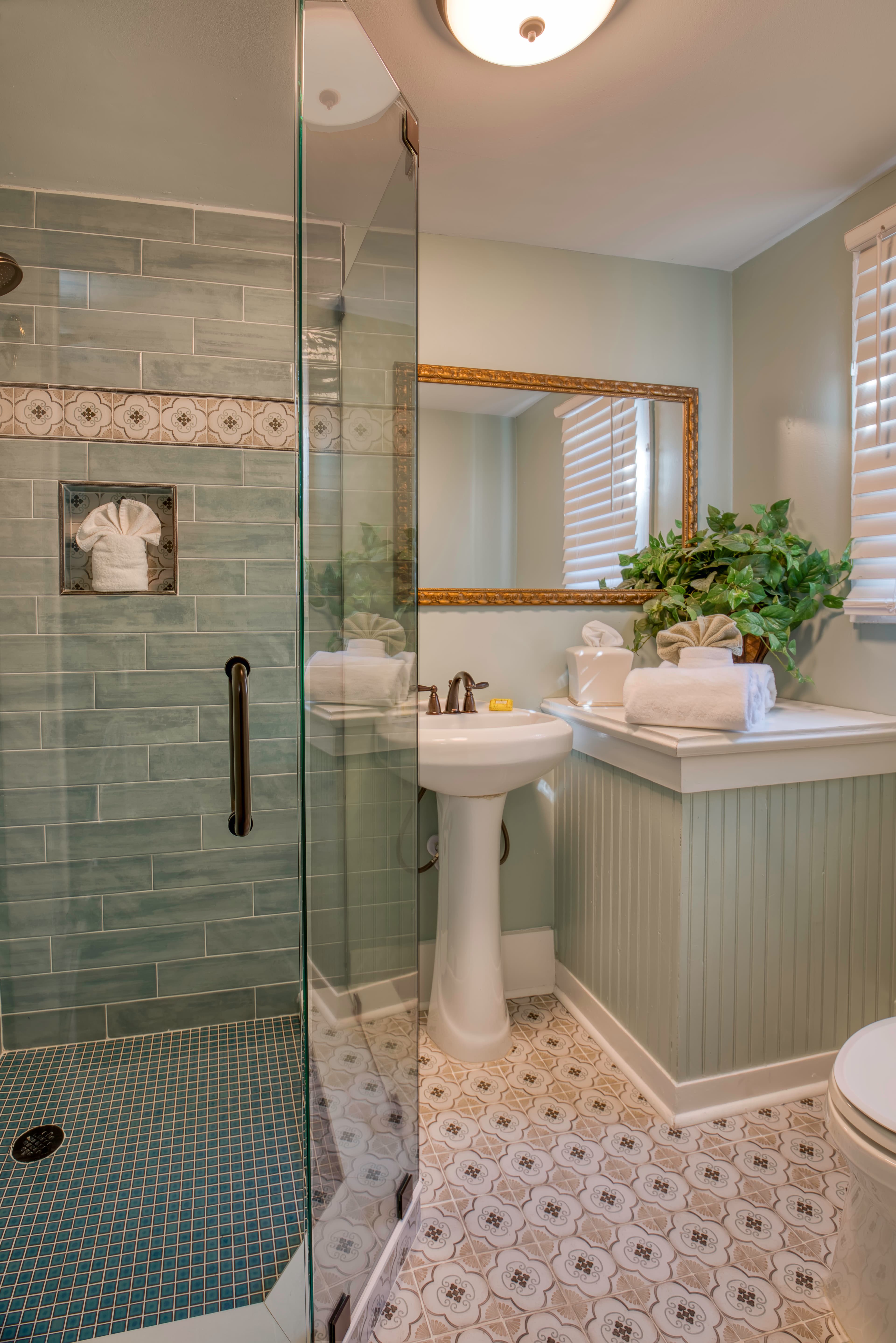 Bathroom in tranquil light blues & white, glass-enclosed tiled shower, white pedestal sink, toilet, and tile floor