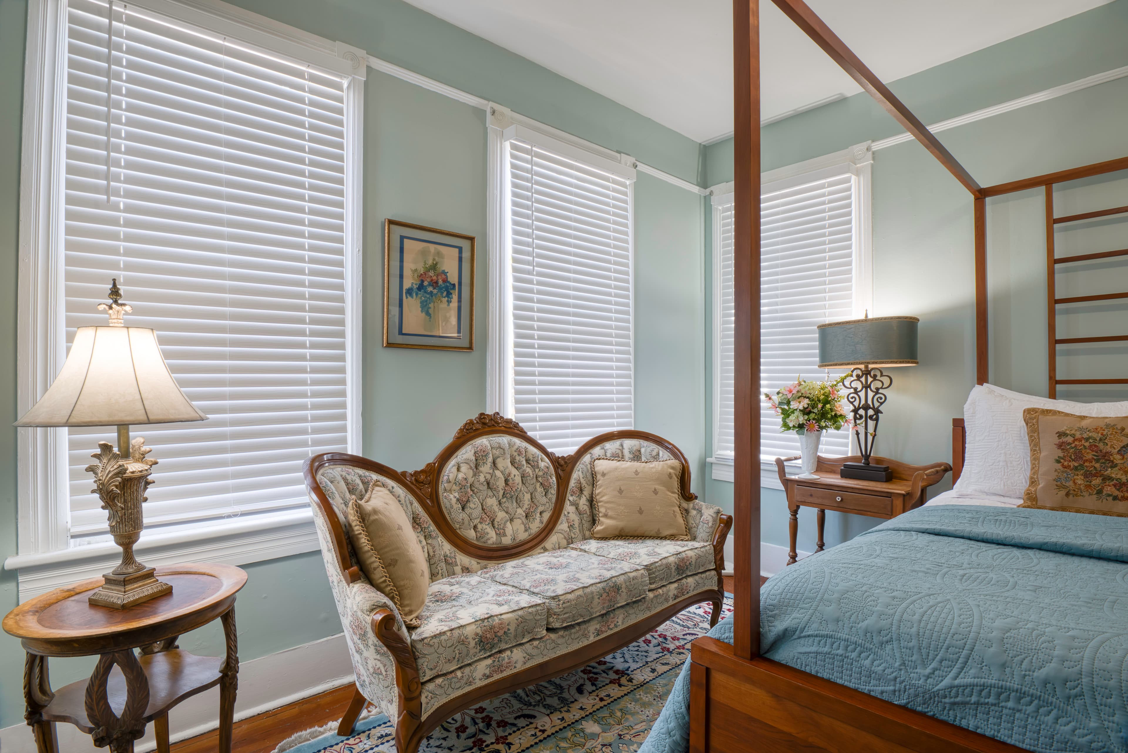 Sitting area in bedroom with Victorian-style couch, oval side table with lamp, and large windows with blinds