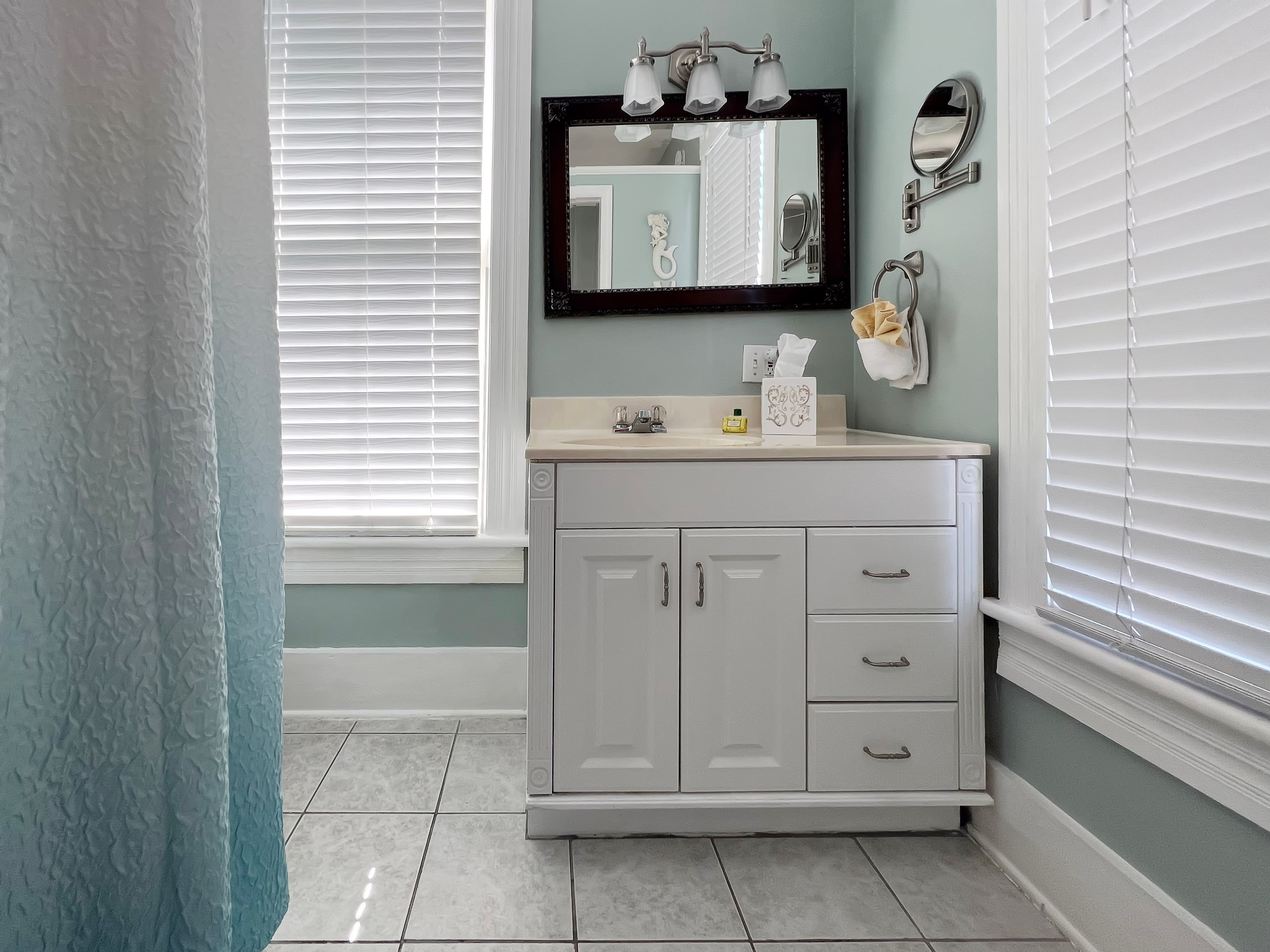 Bathroom in white and shades of blue, white vanity with sink, large windows with blinds, and shower