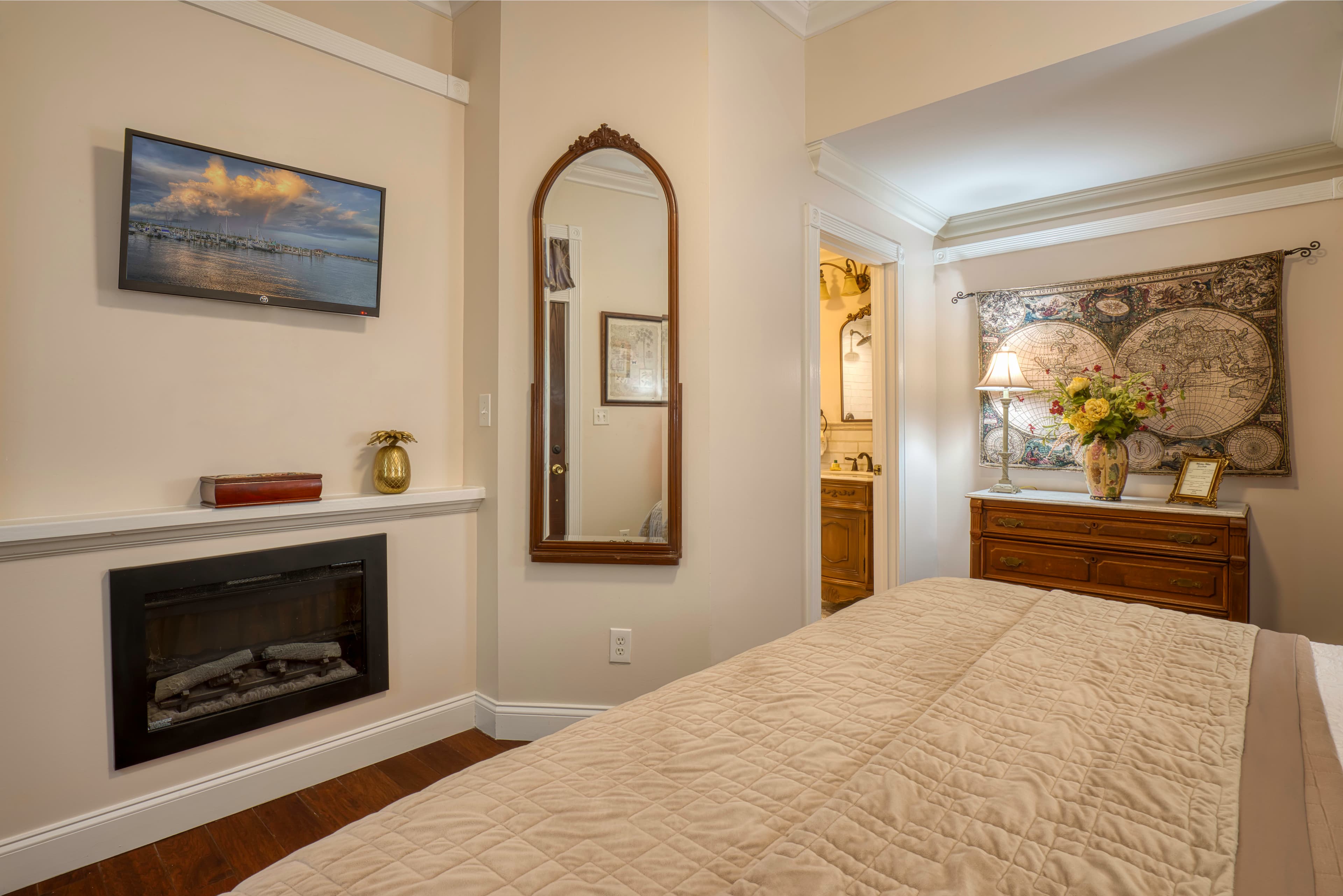 Area opposite foot of bed, fireplace, wall-mounted flat-screen TV, decorative tapestry hanging above wooden dresser, doorway to bathroom