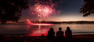 Three people watch colorful fireworks reflecting on the water during twilight. Three people watch colorful fireworks reflecting on the water during twilight.